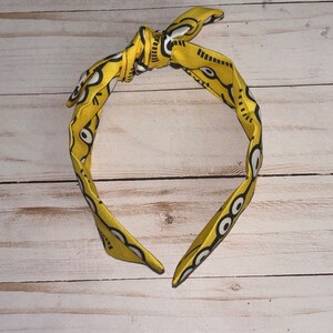 May include: A yellow headband with a black and white geometric pattern. The headband is tied in a knot at the top.