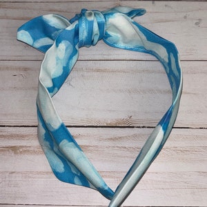 May include: A blue and white tie-dye headband with a knotted bow. The headband is made of fabric and is designed to be worn around the head.