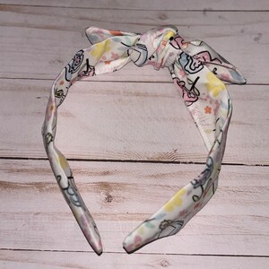 May include: A white headband with a floral pattern and a bow. The headband features a pink, yellow, and blue floral design with small white flowers.