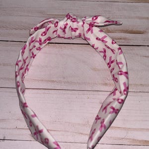 May include: White headband with a pink ribbon pattern. The headband is tied in a bow at the top.