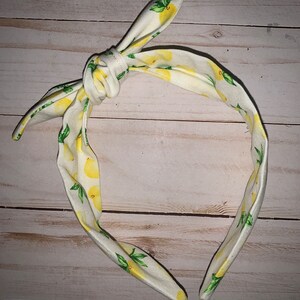 May include: A white headband with a knotted bow, adorned with a repeating pattern of yellow lemons and green leaves. The headband is designed for comfort and style, perfect for adding a touch of summer to any outfit.