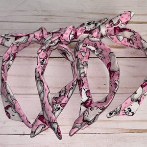 May include: Three pink headbands with a white cat print and pink polka dots. The headbands are tied in a bow.