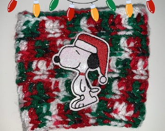 May include: A red, white, and green crocheted coffee cup cozy with a glittery finish. The cozy features an embroidered image of Snoopy wearing a red Santa hat.