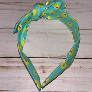 May include: A turquoise headband with a pattern of yellow avocados. The headband is tied in a knot at the top.
