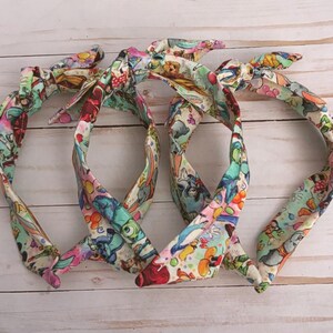 May include: Four colorful fabric headbands with a knotted bow design. The headbands feature a print with cartoon characters in shades of blue, pink, green, and red. The headbands are arranged on a light wood-grain surface.