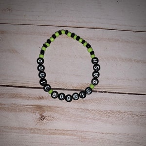 May include: A beaded bracelet with black and neon green beads. The bracelet spells out "BOOGIE BASH" in black letter beads. The bracelet is on a light wood grain surface.
