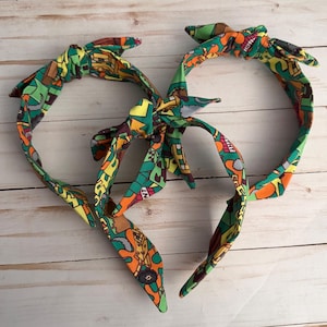 May include: Two fabric headbands featuring a vibrant, multi-colored graphic print with green, orange, yellow, and brown geometric shapes and patterns. Each headband is tied at the top with a decorative knot, creating a bow-like appearance.