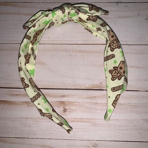 May include: A fabric headband with a bow, featuring a repeating pattern of a cartoon character and green stars on a light green background. The character's name, "GROOT", is printed on the headband.