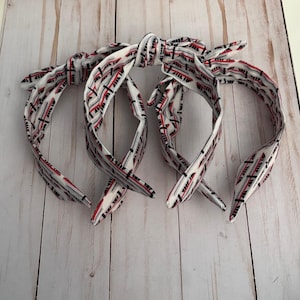 May include: Four white headbands with a red and black geometric pattern. Each headband is tied in a knot at the top, with the ends forming a bow. The headbands are made of fabric and are designed to be worn on the head.