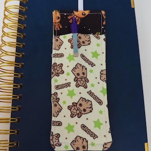May include: A navy blue spiral-bound notebook with a fabric pen holder featuring a repeating pattern of the character Groot from Guardians of the Galaxy. The pen holder is attached to the notebook with a white elastic band and contains a purple pen.