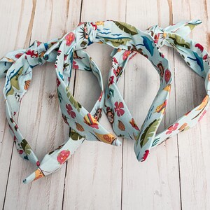 May include: Three light blue headbands with a tropical print featuring flowers, leaves, and sea creatures. The headbands are tied in a knot at the top, with the ends of the fabric extending out. The headbands are made of a soft, flexible material.