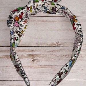 May include: A white headband with a colorful print of cartoon characters and stars. The headband is tied in a knot at the top.