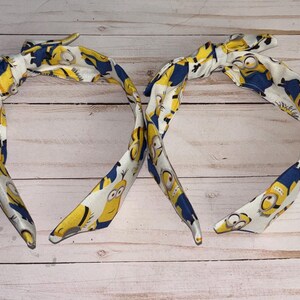 May include: Two yellow and blue headbands with a cartoon minion pattern. The headbands are tied in a bow at the top.