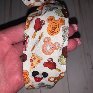 May include: A white headband with a colorful print of Mickey Mouse and various Disney snacks, including pretzels, popcorn, pizza, ice cream, and cupcakes.
