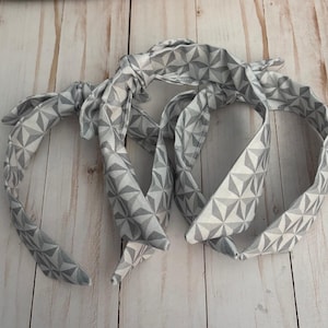 May include: Three gray and white geometric patterned headbands with tied bows. The headbands are made of fabric and have a repeating triangular design. The headbands are laying on a light wood surface.