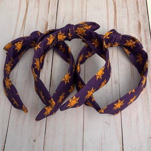 May include: Three purple headbands with a repeating yellow sun pattern. Each headband is tied in a bow at the top. The headbands are made of fabric and are displayed on a light wood surface.