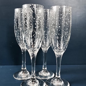 Op de afbeelding: Vier heldere champagneglazen met witte gespikkelde patronen. De glazen hebben een hoge, slanke vorm met een ronde voet en een lange steel. De achtergrond is donkerblauw.