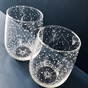 May include: Two clear stemless wine glasses with a constellation design. The glasses are decorated with white dots and lines, creating a starry night effect. A star is at the bottom of each glass. The glasses sit on a dark blue surface.