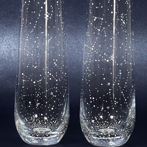 May include: Two clear champagne flutes with a constellation-like pattern of white dots. The glasses have a rounded base and a tall, slender shape. The white dots vary in size and density, creating a starry effect.