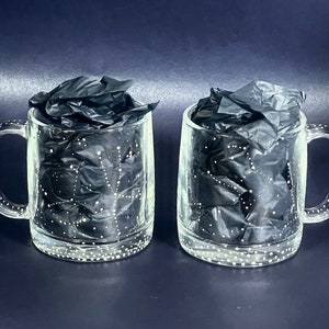 Könnte beinhalten: Zwei durchsichtige Glasbecher mit weißen Punkten und schwarzem Seidenpapier im Inneren. Die Becher haben Henkel.