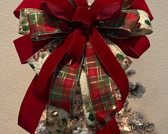 May include: A red velvet and plaid Christmas tree topper bow with gold accents. The bow is tied with a large, red velvet ribbon and features a plaid ribbon with gold accents.