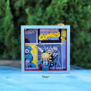 May include: A blue wooden frame with a comic book image inside. The comic book image features a superhero with a hammer. The frame has the word "Thor" printed below it.