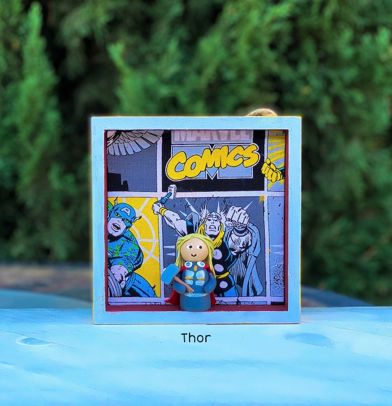 May include: A blue wooden frame with a comic book image inside. The comic book image features a superhero with a hammer. The frame has the word "Thor" printed below it.