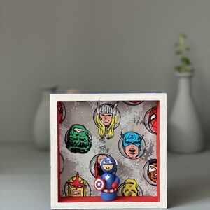 May include: A small framed shadow box with a red and white border. The box contains a small clay figurine of Captain America holding his shield. The background of the box is a grey fabric with a pattern of comic book superhero faces.