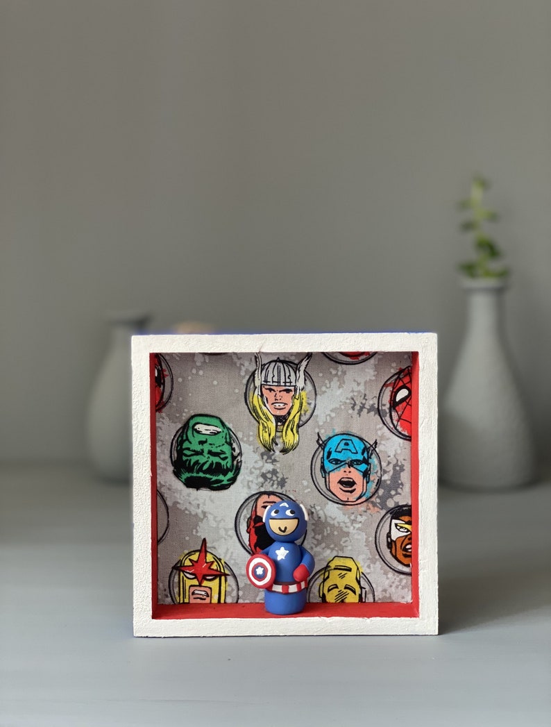May include: A small framed shadow box with a red and white border. The box contains a small clay figurine of Captain America holding his shield. The background of the box is a grey fabric with a pattern of comic book superhero faces.