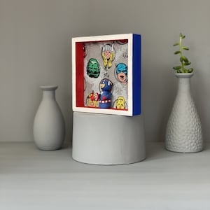 May include: A framed shadow box with a red, white and blue border. Inside the box is a small figurine of Captain America standing in front of a comic book background featuring various superhero faces.