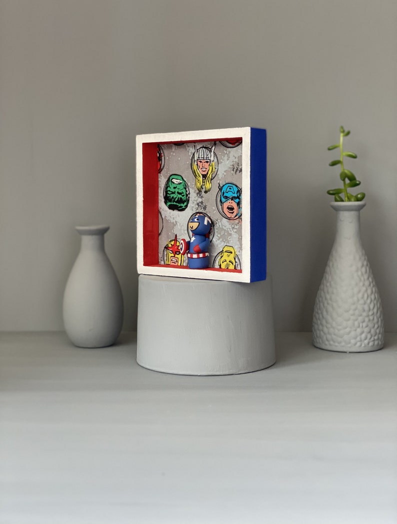 May include: A framed shadow box with a red, white and blue border. Inside the box is a small figurine of Captain America standing in front of a comic book background featuring various superhero faces.
