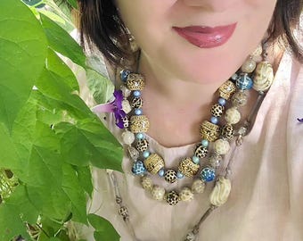 May include: A multi-strand beaded necklace featuring a mix of textured tan, brown, and blue beads, including patterned spherical beads and smooth light blue accents, worn over a plain light-colored top with a soft, neutral fabric texture.