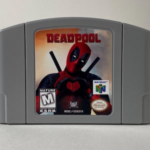 May include: A gray Nintendo 64 game cartridge with a red, black, and white label featuring Deadpool making a heart shape with his hands. The label also includes the game title "DEADPOOL", a mature rating label, and the Nintendo 64 logo.