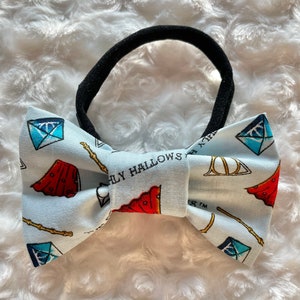 May include: A light blue headband with a bow featuring a Harry Potter themed print. The print includes the Deathly Hallows symbol, wands, and a red cloak.