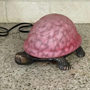 May include: A decorative turtle-shaped oil warmer. The turtle has a bronze-colored base with a pink, textured shell. The warmer is designed to hold essential oils and is a unique home decor item.