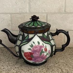 May include: A stained glass teapot lamp featuring a floral motif with pink and red roses on a white background. The lamp has a dark metal frame, a curved handle, a spout, and a lid topped with a small metal rose finial. A black power cord extends from the base.