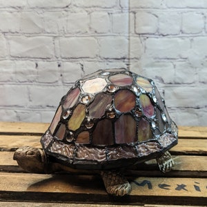 May include: A decorative turtle figurine with a stained glass shell. The shell features a mosaic of purple, tan, and clear glass pieces. The turtle's body and head are a textured brown color, and it sits on a wooden surface.