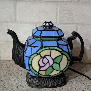 May include: A stained glass teapot lamp with a black handle, spout, and base. The lamp features a blue, green, and white geometric design with a pink rose in the center. The lamp is plugged into an electrical outlet.