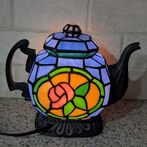 Pode incluir: Uma l&acirc;mpada decorativa em forma de bule de vitral. A l&acirc;mpada tem uma base e uma al&ccedil;a pretas. O design mostra um padr&atilde;o colorido de vitral com tons de azul, verde, laranja e rosa, formando um motivo floral. A l&acirc;mpada est&aacute; ligada e iluminada.