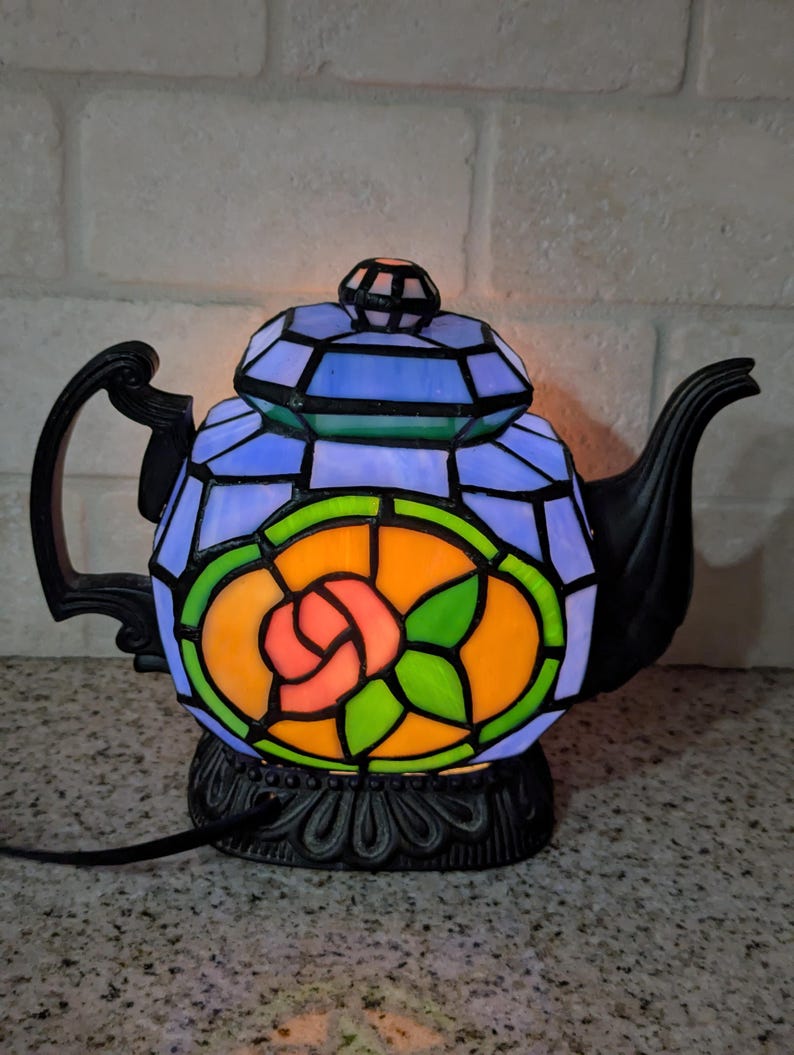 Pode incluir: Uma l&acirc;mpada decorativa em forma de bule de vitral. A l&acirc;mpada tem uma base e uma al&ccedil;a pretas. O design mostra um padr&atilde;o colorido de vitral com tons de azul, verde, laranja e rosa, formando um motivo floral. A l&acirc;mpada est&aacute; ligada e iluminada.