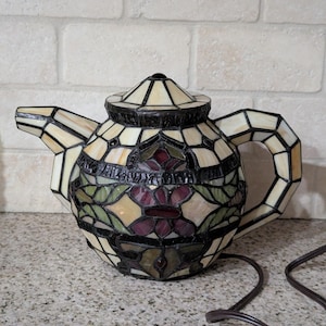 May include: A stained glass teapot lamp with a floral design. The lamp features a cream-colored background with green, red, and purple floral accents. The handle, spout, and lid are also made of stained glass.