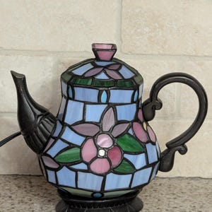 May include: A decorative stained glass teapot lamp with a black handle, spout, and base. The lamp features a floral design in shades of blue, pink, green, and purple. The lamp is designed to be plugged into an electrical outlet.