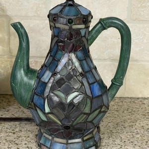 May include: A decorative stained glass teapot lamp with a green handle and spout. The lamp features a mosaic design in shades of blue, green, and purple. The base and lid are also stained glass, creating a unique lighting fixture.