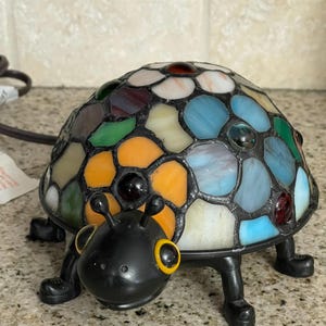 May include: A colorful stained glass turtle lamp with a black base and legs. The turtle's shell is made of various colored glass pieces, including orange, blue, green, and white. The turtle's head is black with yellow accents.
