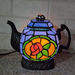 Pode incluir: L&acirc;mpada decorativa em forma de bule com design de vitral. A l&acirc;mpada tem uma base e uma al&ccedil;a pretas, com um design colorido de rosa no centro. A parte superior &eacute; azul e a base &eacute; preta.