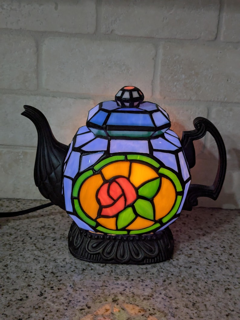 Pode incluir: L&acirc;mpada decorativa em forma de bule com design de vitral. A l&acirc;mpada tem uma base e uma al&ccedil;a pretas, com um design colorido de rosa no centro. A parte superior &eacute; azul e a base &eacute; preta.