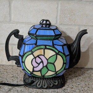 Pode incluir: L&acirc;mpada de bule de vitral com al&ccedil;a e bico pretos. A l&acirc;mpada apresenta um design geom&eacute;trico azul e verde com uma rosa rosa no centro. A base &eacute; preta e ornamentada.
