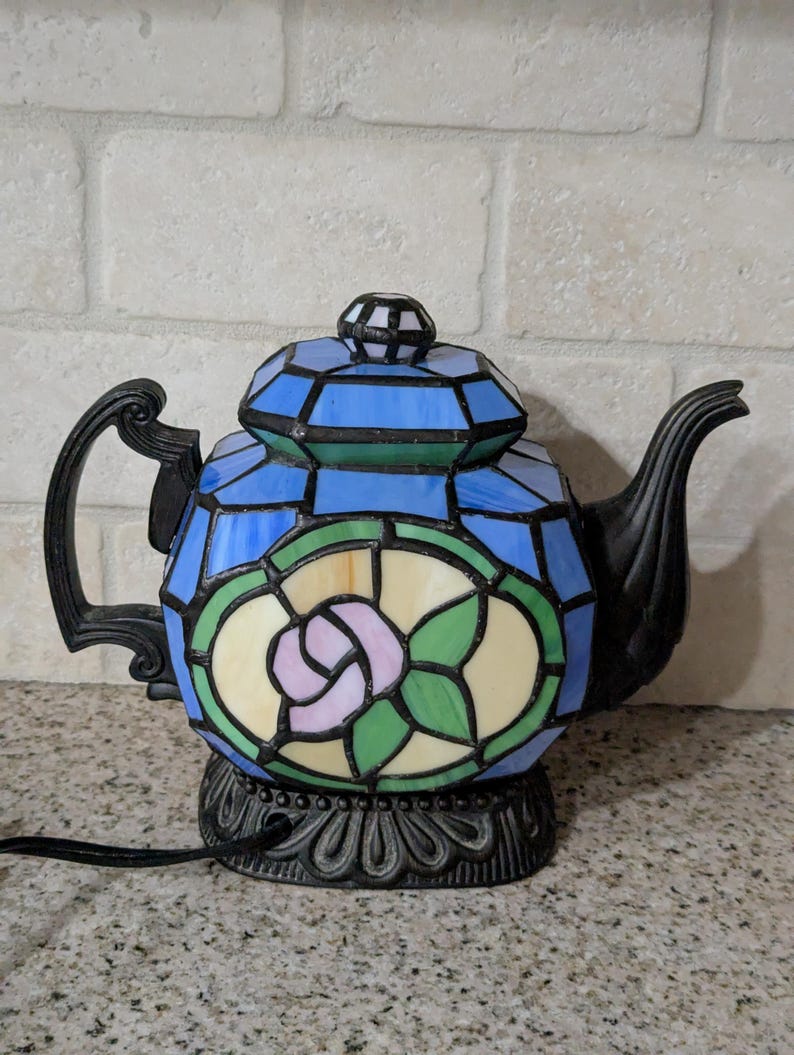 Pode incluir: L&acirc;mpada de bule de vitral com al&ccedil;a e bico pretos. A l&acirc;mpada apresenta um design geom&eacute;trico azul e verde com uma rosa rosa no centro. A base &eacute; preta e ornamentada.