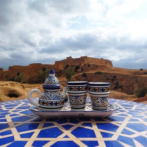 Peut inclure: Un service à thé en céramique composé d'une petite théière et de deux tasses assorties disposées sur un plateau ovale. Les pièces sont blanches avec des motifs géométriques peints à la main en bleu, jaune et noir sur une table carrelée.