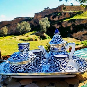 Peut inclure: Un service à thé en métal argenté comprenant une théière, un sucrier avec couvercle et trois petites tasses, ornés de motifs géométriques bleus et blancs. L'ensemble est disposé sur un plateau de service rectangulaire en métal argenté.